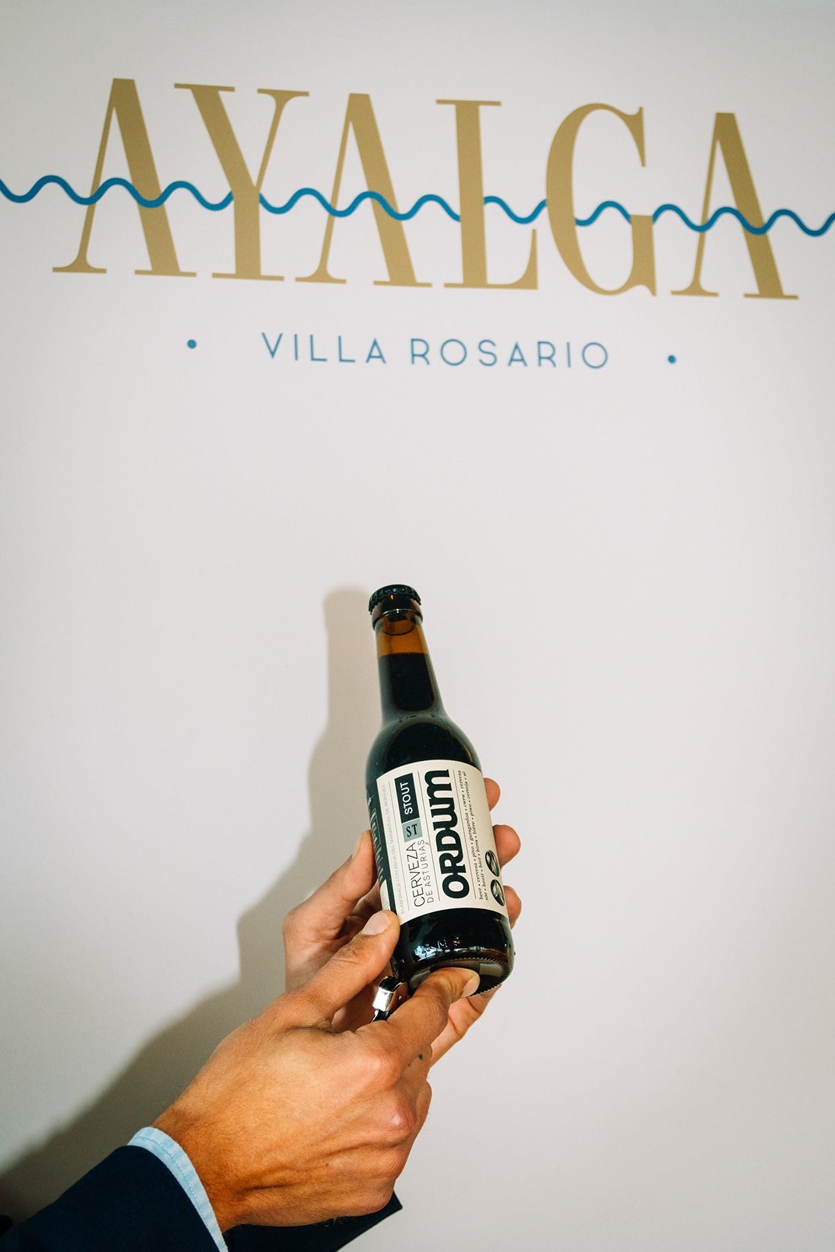 Mano sosteniendo una botella de cerveza oscura frente a un cartel de Ayalga Villa Rosario.