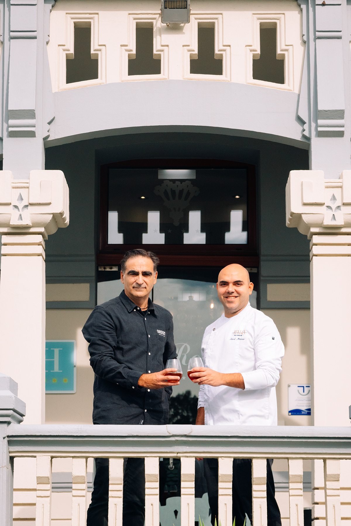 Ayalga x Ordum: dos hombres, uno con chaqueta de cocinero, posando con copas en la entrada del edificio