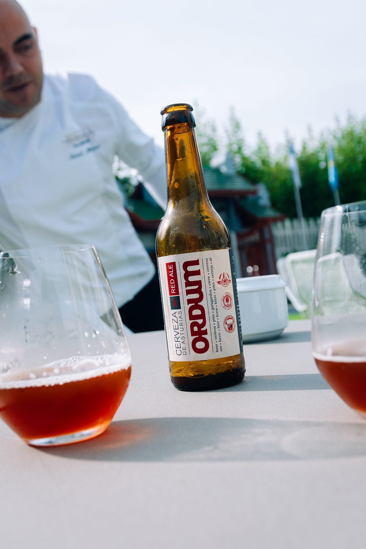 Ayalga x Ordum: botella de Ordum y copas de cerveza en una mesa con un cocinero al fondo