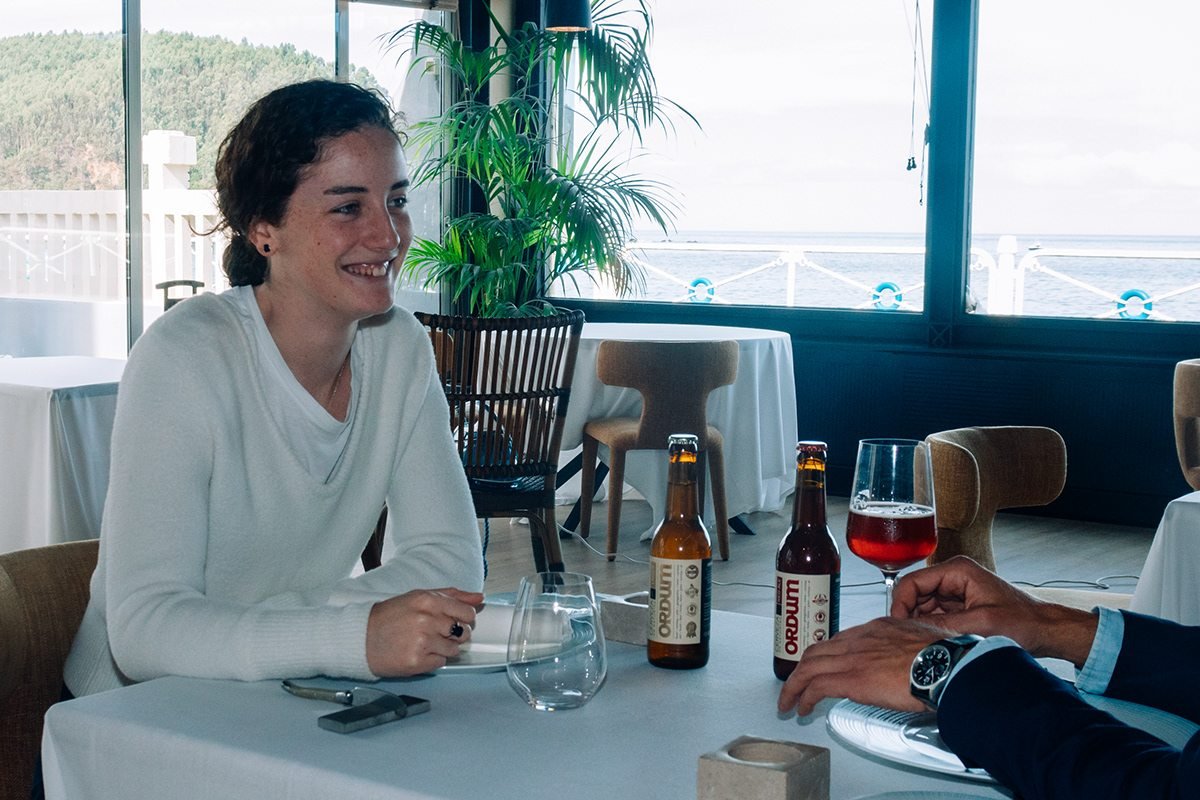 Ayalga x Ordum: mujer sonriente en una mesa con cerveza Ordum junto a un ventanal
