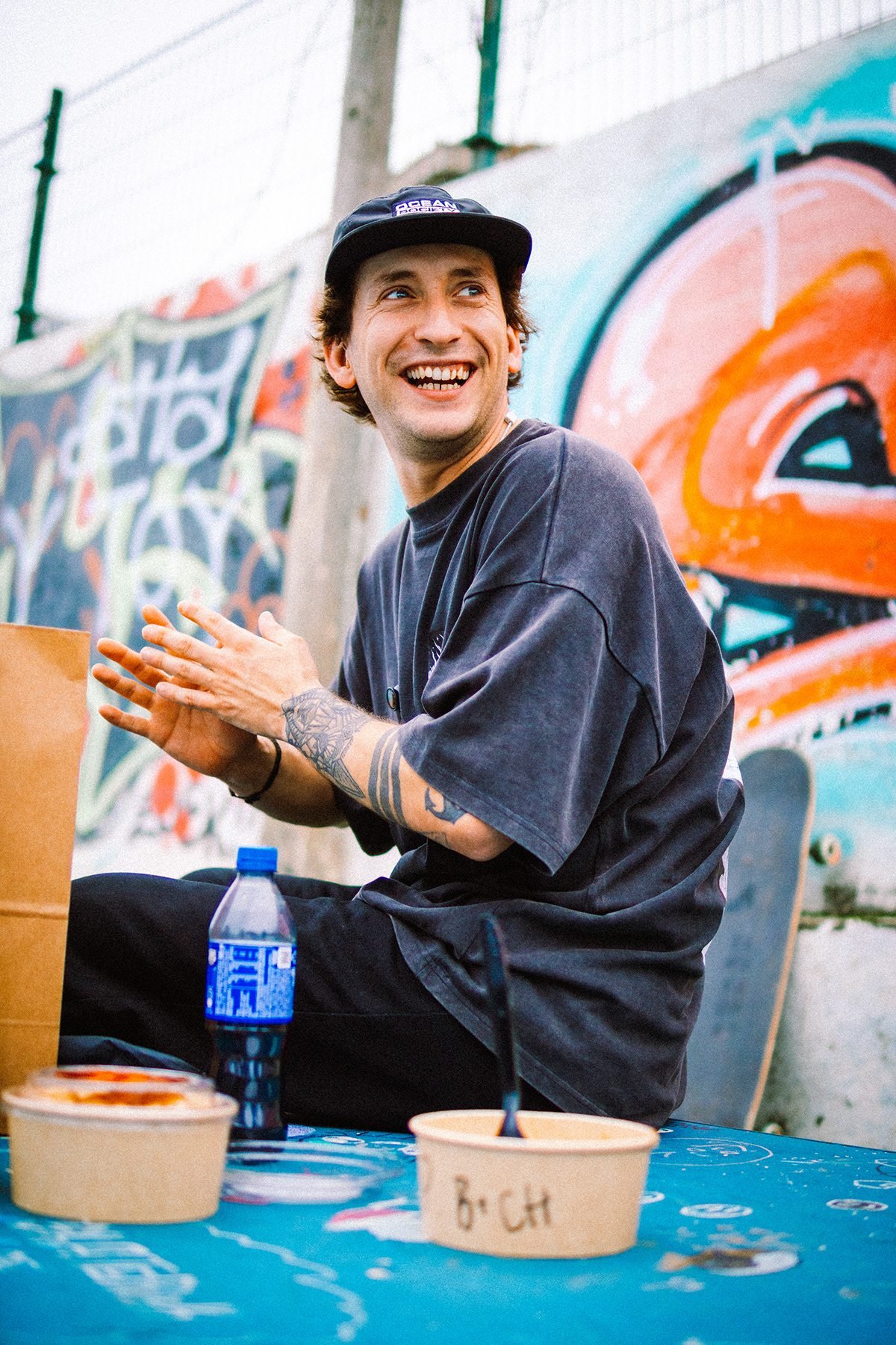 Hombre con gorra sonriendo sentado junto a un mural de graffiti, con envases de comida en primer plano.
