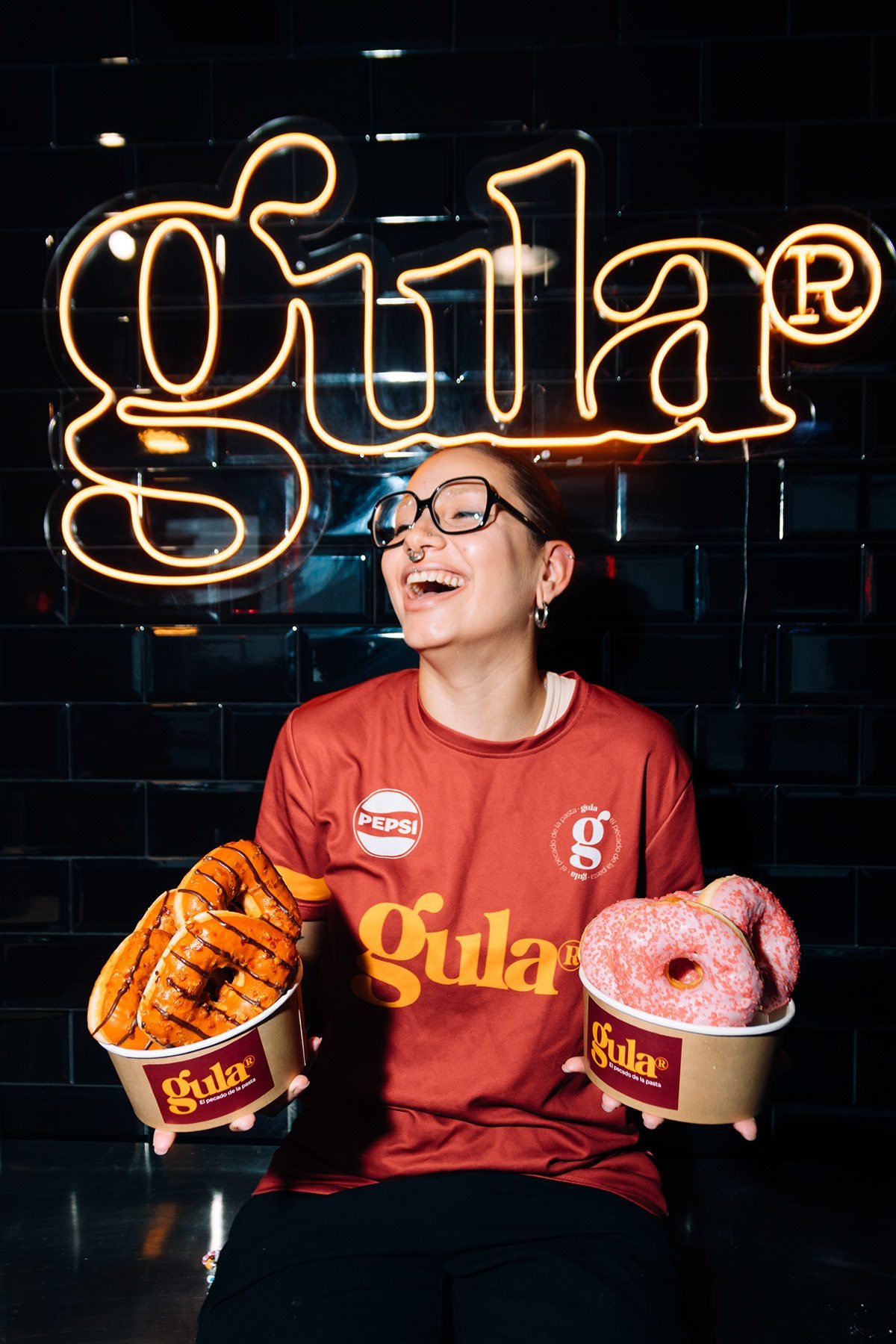 Gula: mujer con camiseta de Gula sosteniendo dos boles de dulces bajo el neón de la marca