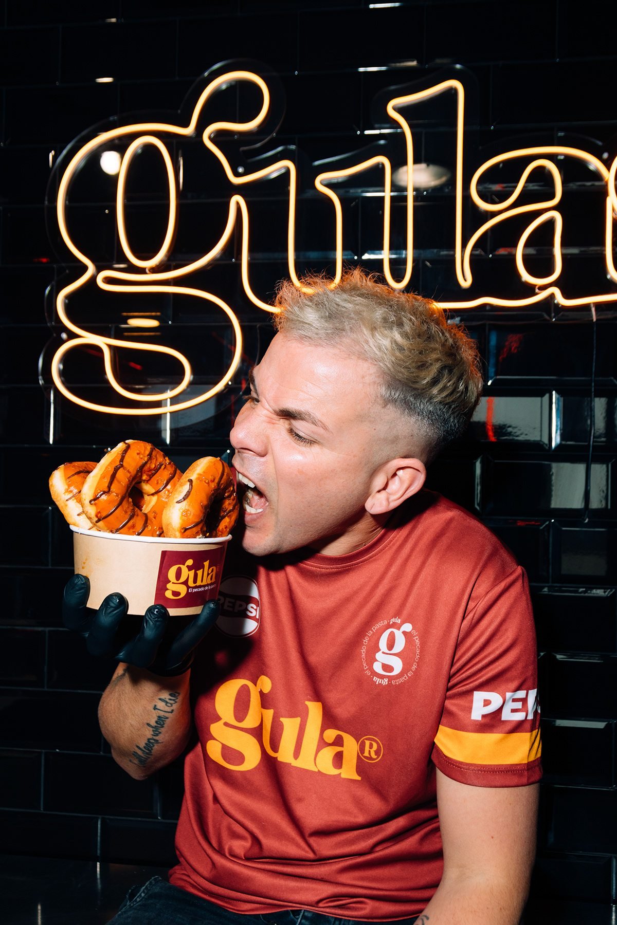 Gula: hombre con camiseta de Gula mordiendo un dulce bajo el neón de la marca