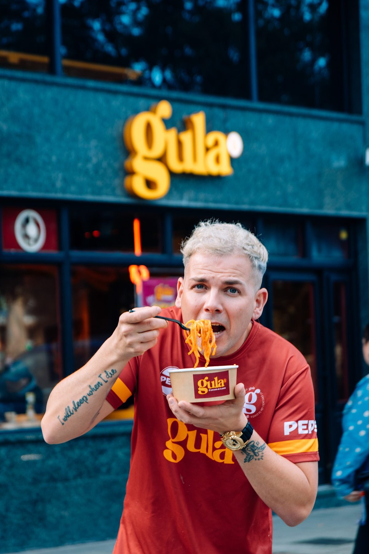 Gula: hombre con camiseta de Gula comiendo pasta frente al rótulo del local