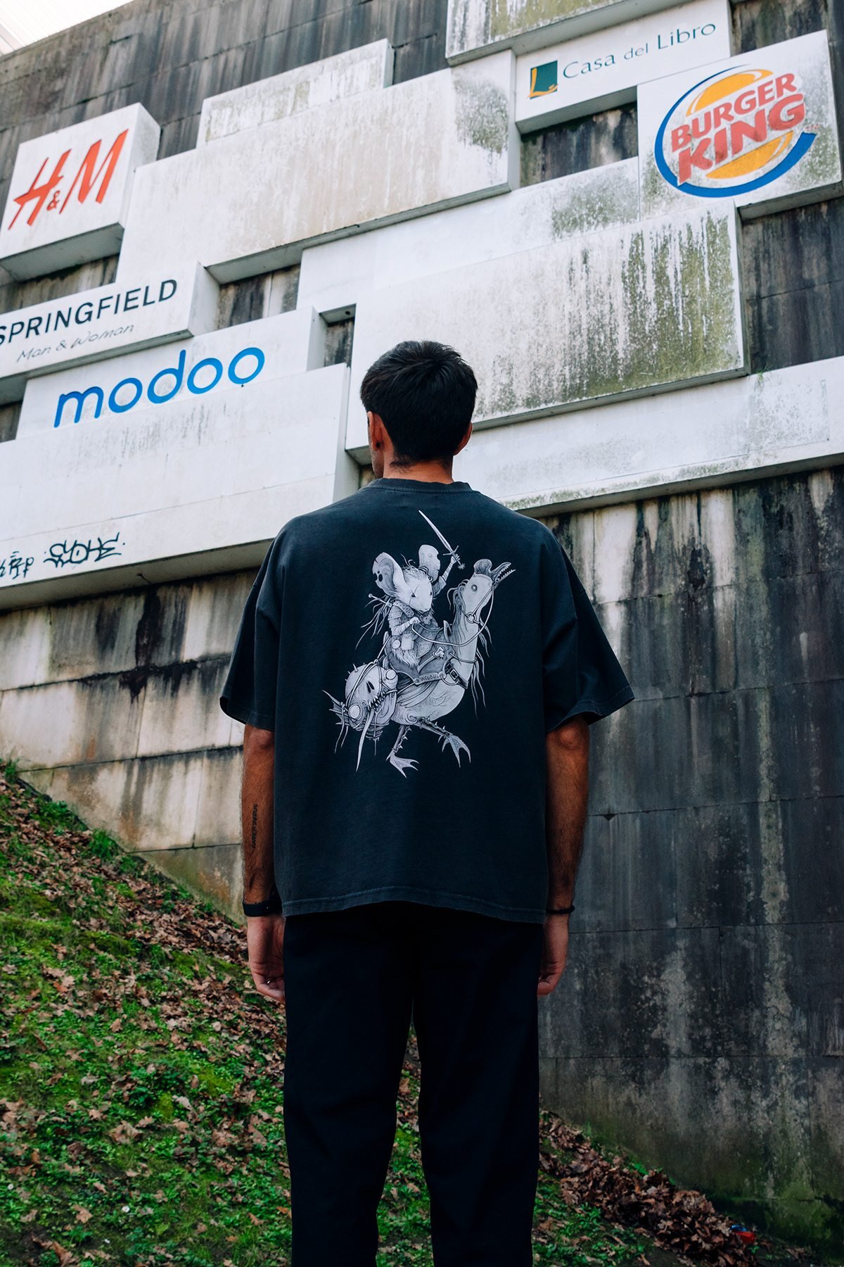 Incubu Clothing: modelo de espaldas con camiseta estampada frente a un edificio con rótulos comerciales
