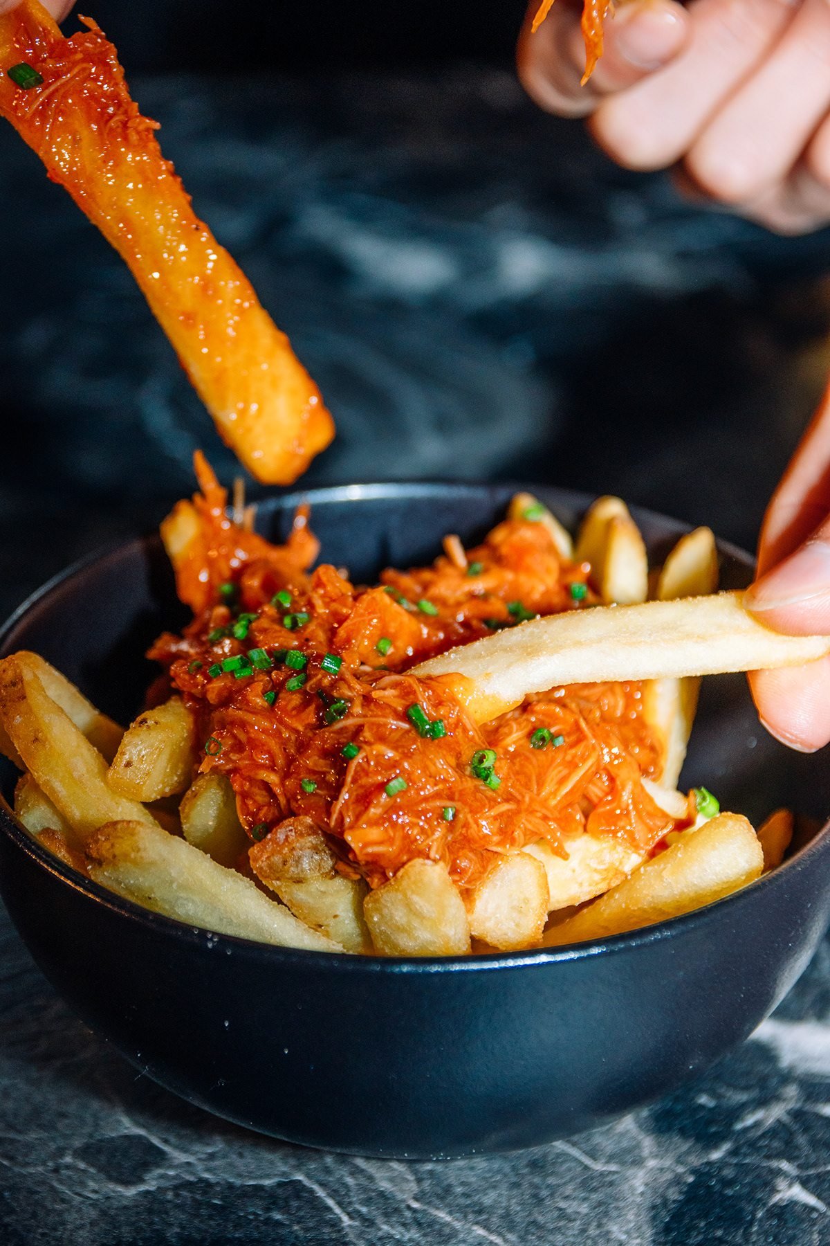 Mr Kong: mano mojando una patata frita en un bol de patatas con carne desmechada