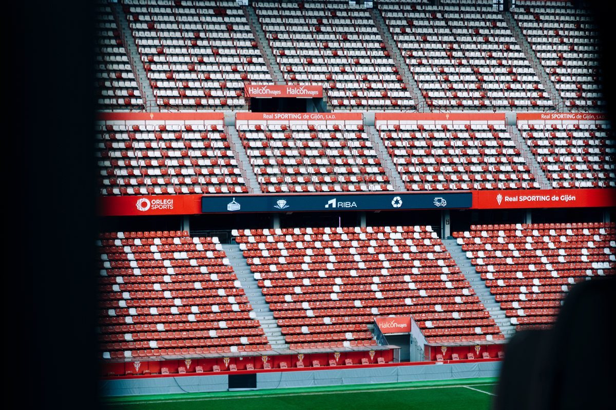 Gradas de un estadio de fútbol con asientos rojos y blancos y el césped al fondo.
