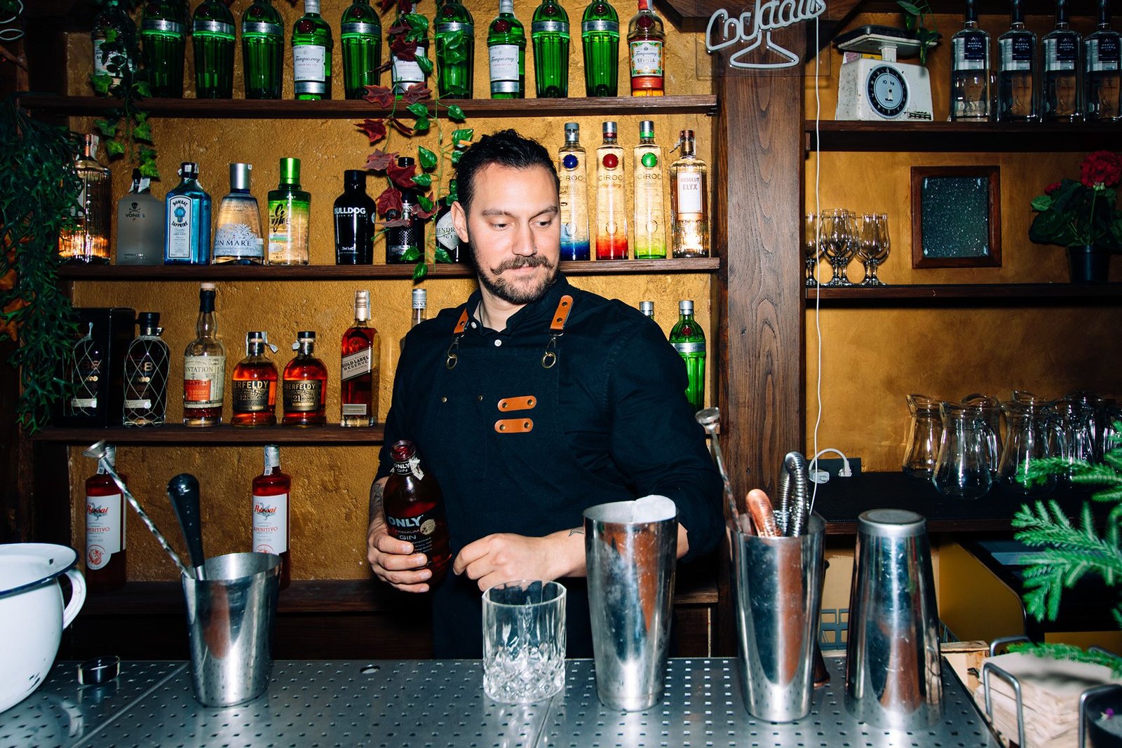 Barista prepara una bebida en la barra con botellas y utensilios al fondo.