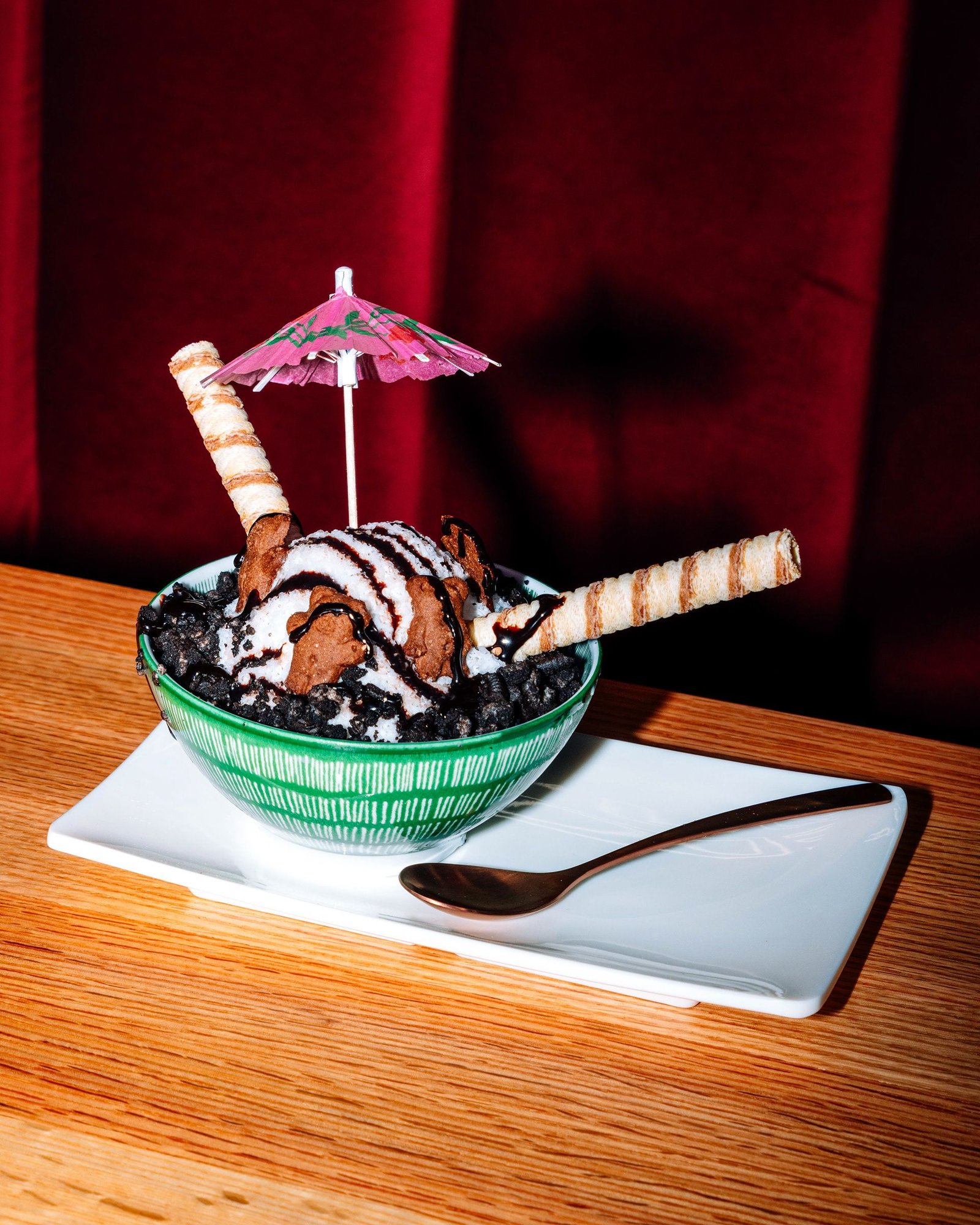 Helado con galleta de chocolate, sirope y barquillos, decorado con una sombrilla de papel, en un bol verde sobre una mesa de madera.