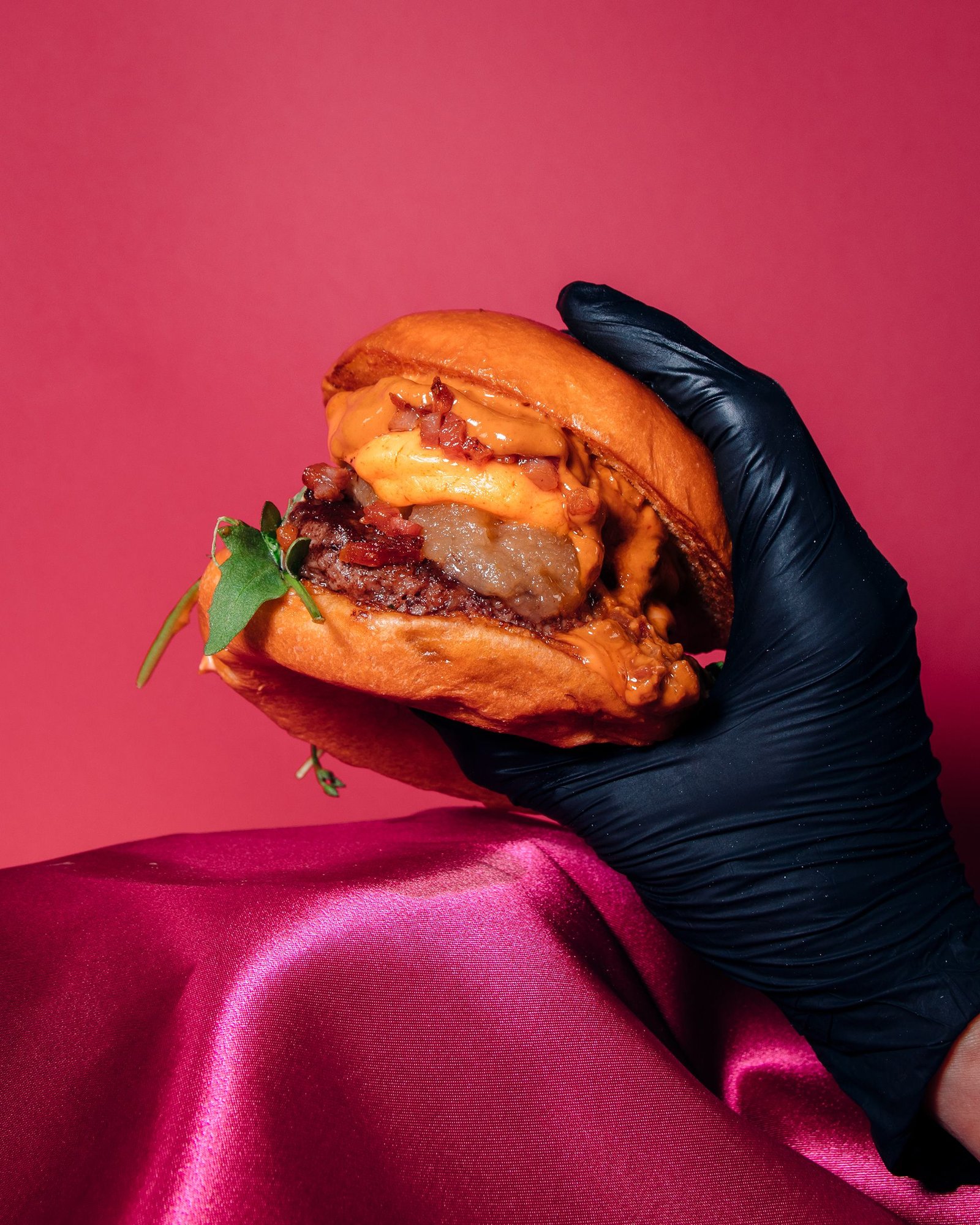Mano enguantada sostiene una hamburguesa sobre una mesa rosa.