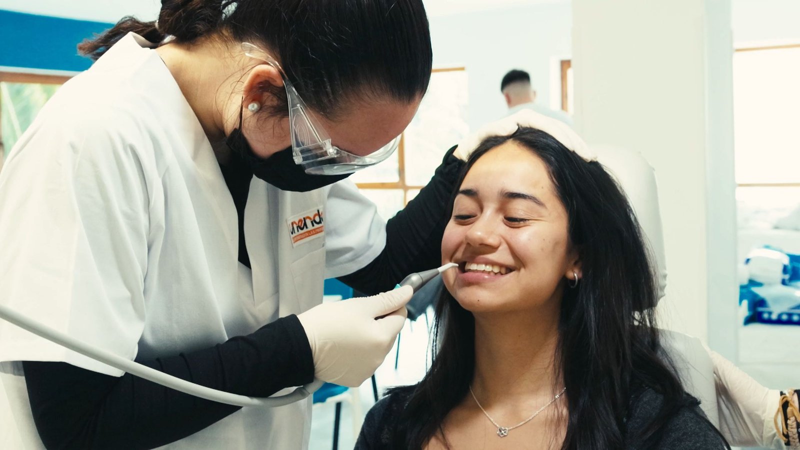 Alumna con bata, mascarilla y gafas de protección usa un instrumento en la boca de una compañera sonriente.