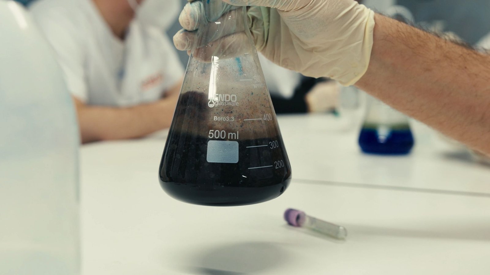 Mano con guante sostiene un matraz con líquido oscuro en una mesa de laboratorio.