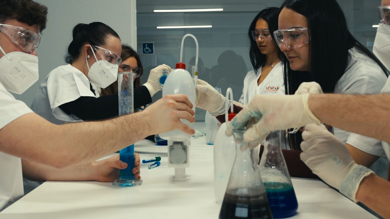 Alumnos con bata, gafas y guantes trabajan con matraces y probetas.