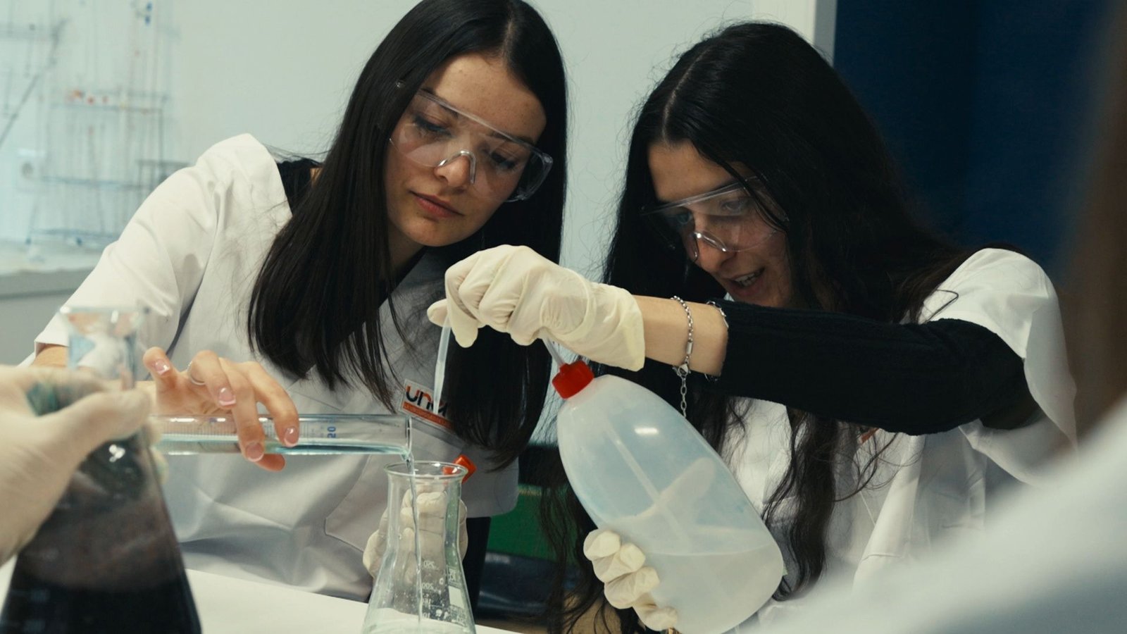 Dos alumnas con bata y gafas de protección trabajan con matraces.