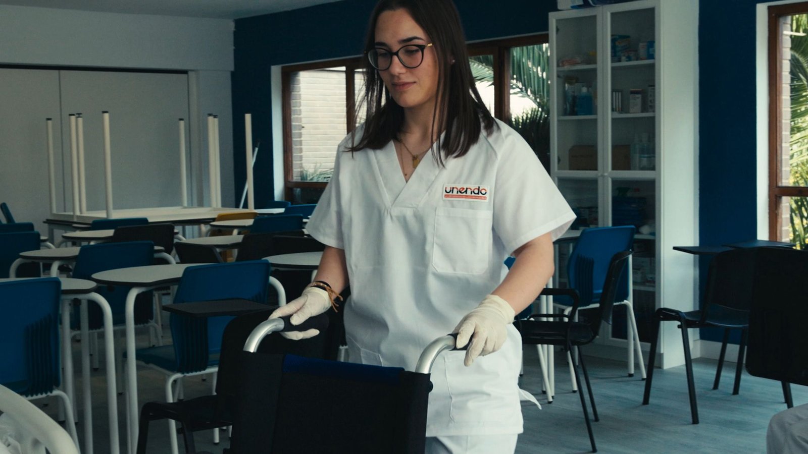 Alumna con bata y guantes empuja una silla de ruedas en un aula.