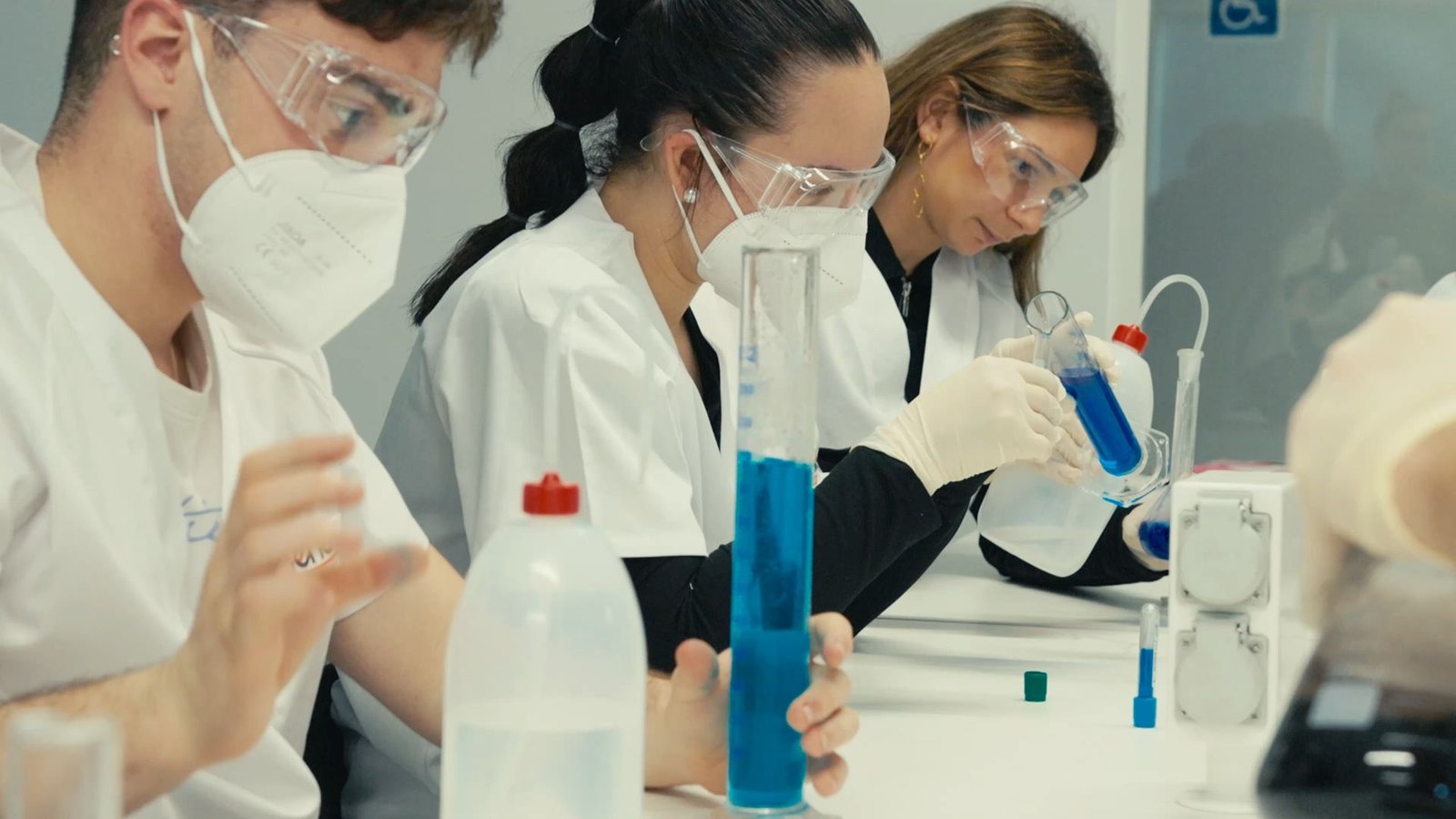 Alumnos con bata y gafas manipulan probetas con líquido azul.
