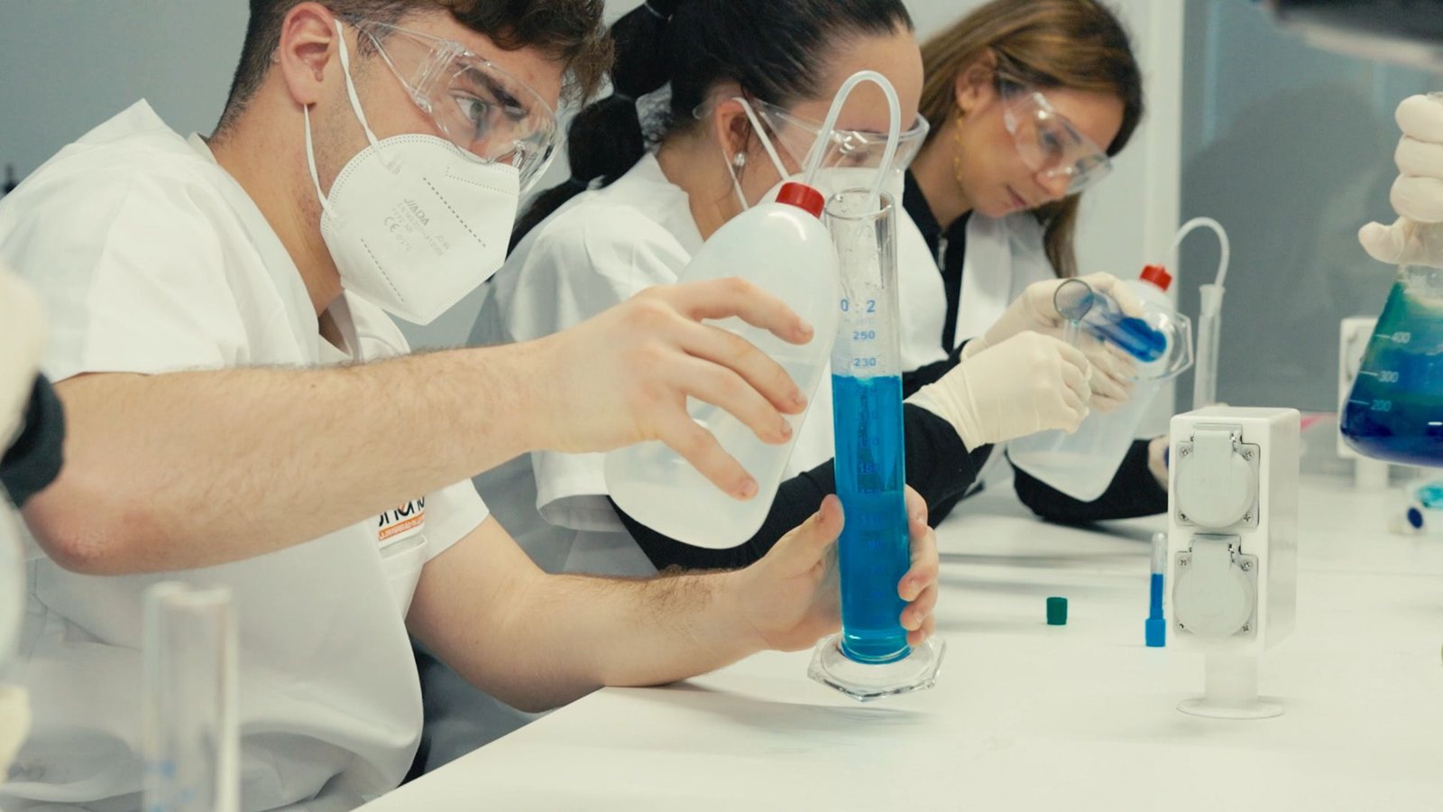 Alumnos con bata, mascarilla y gafas miden líquidos en el laboratorio.
