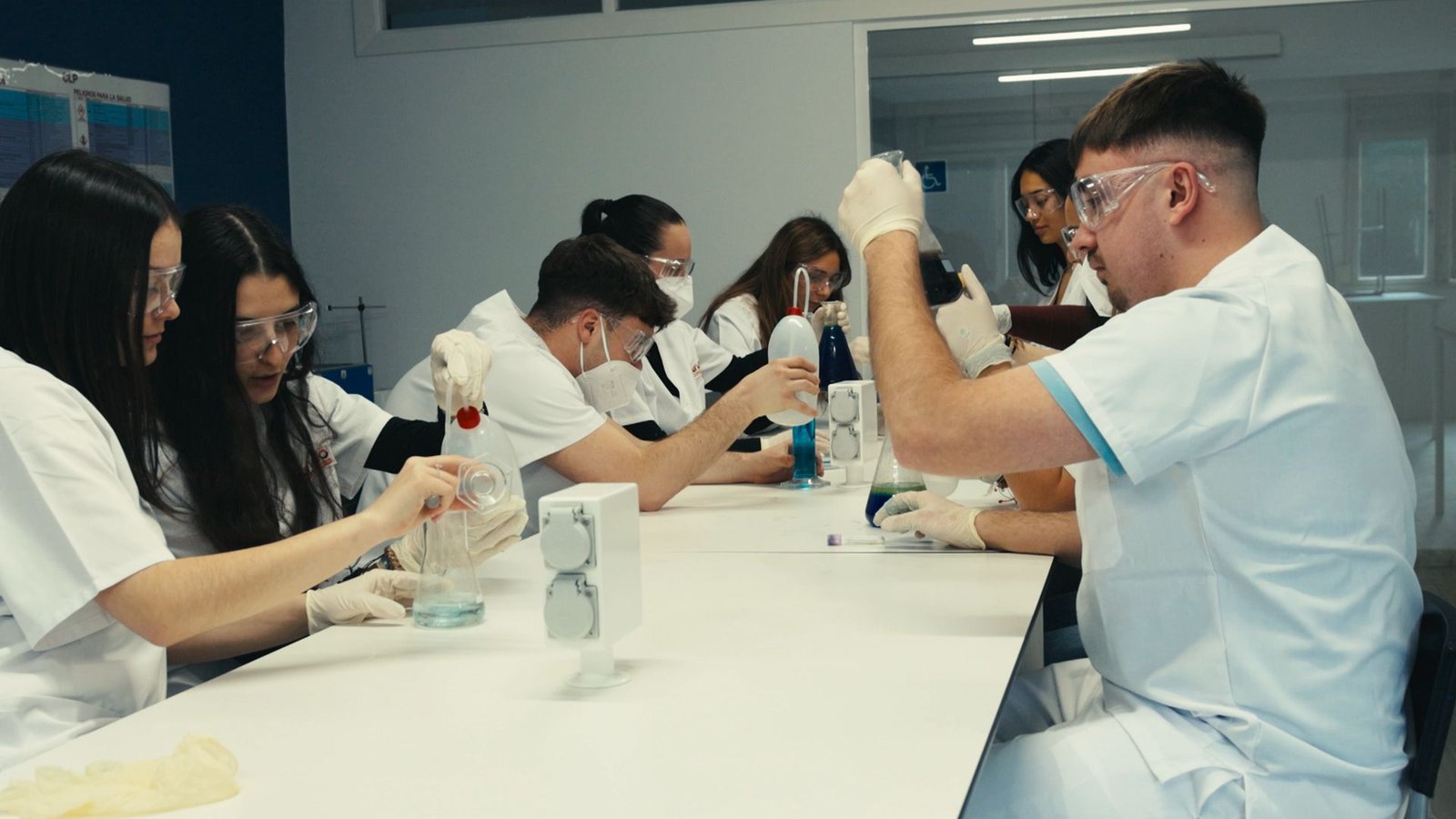 Grupo de alumnos con bata y gafas trabaja en las mesas del laboratorio.